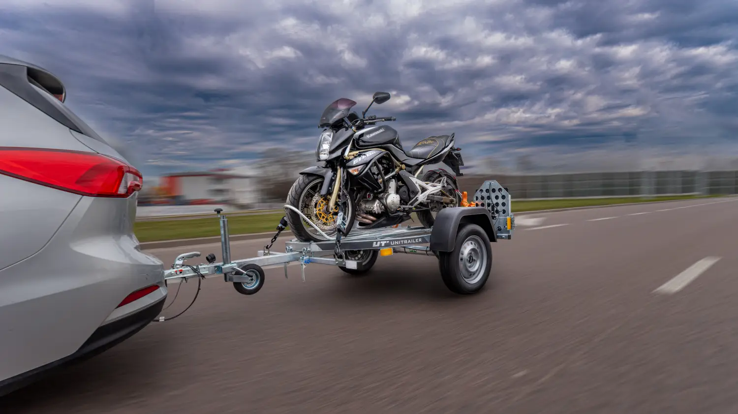 Stříbrný osobní automobil táhnoucí jednonápravový přívěs na motocykly Unitrailer po asfaltové cestě, s upevněným černým motocyklem Kawasaki.