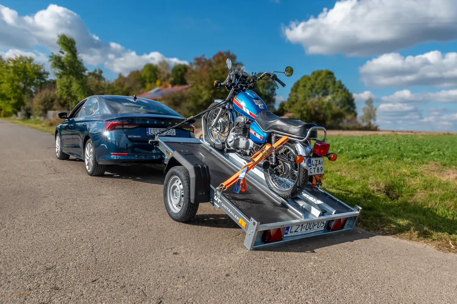 Tmavě modrá Škoda Octavia na venkovské cestě se sklopným přívěsem na motocykly Unitrailer, na kterém je popruhy upevněn klasický modrý motocykl Honda.
