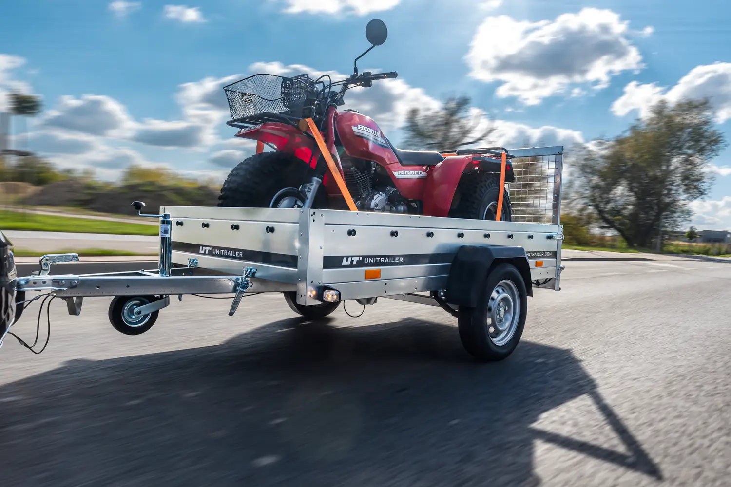 Jednonápravový valníkový přívěs Unitrailer v pohybu, převážející červenou tříkolku Honda s košíkem na řídítkách, zajištěnou oranžovými upínacími popruhy.