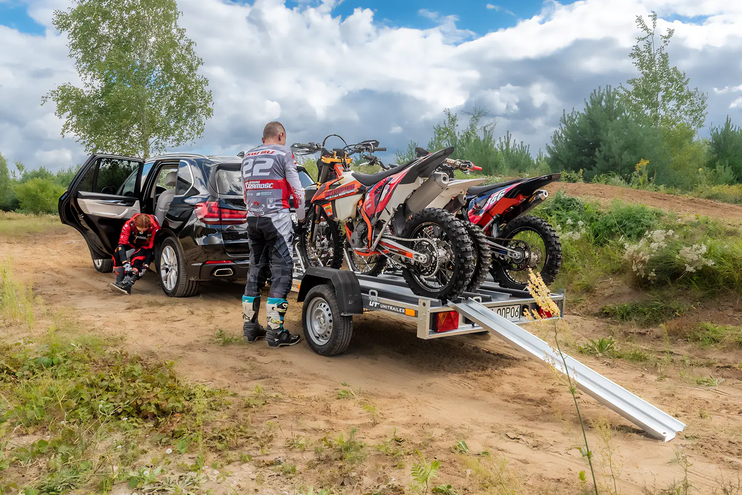 Černý SUV zaparkovaný na terénní trati s přívěsem na motocykly Unitrailer naloženým dvěma motokrosovými stroji, zatímco se dva jezdci v ochranné výstroji připravují vedle nájezdové rampy.