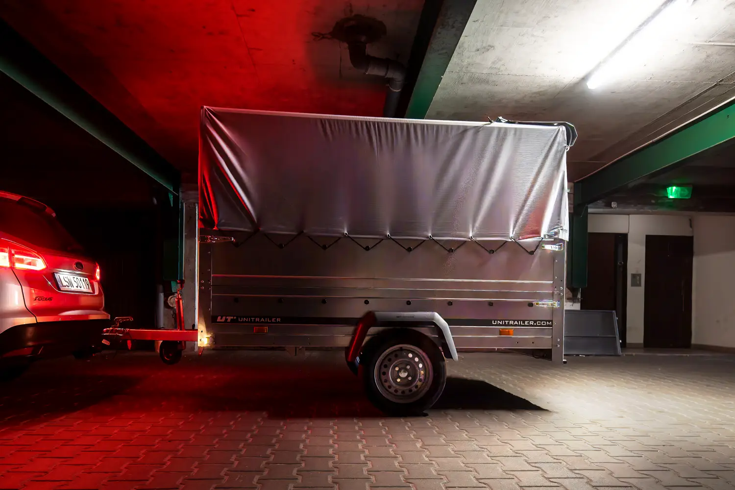 Nákladní přívěs Unitrailer se šedou plachtou, připojený k autu ve slabě osvětlené podzemní garáži.