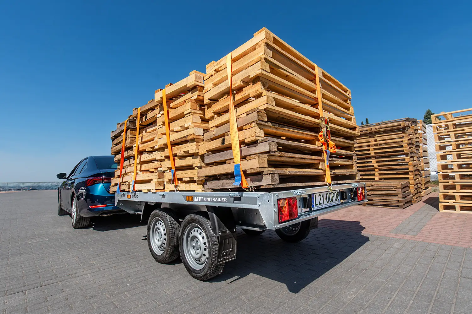 Dvounápravový nákladní přívěs Unitrailer připojený k tmavému autu, naložený vysokou hromadou dřevěných palet zajištěných upínacími pásy na firemním dvoře.