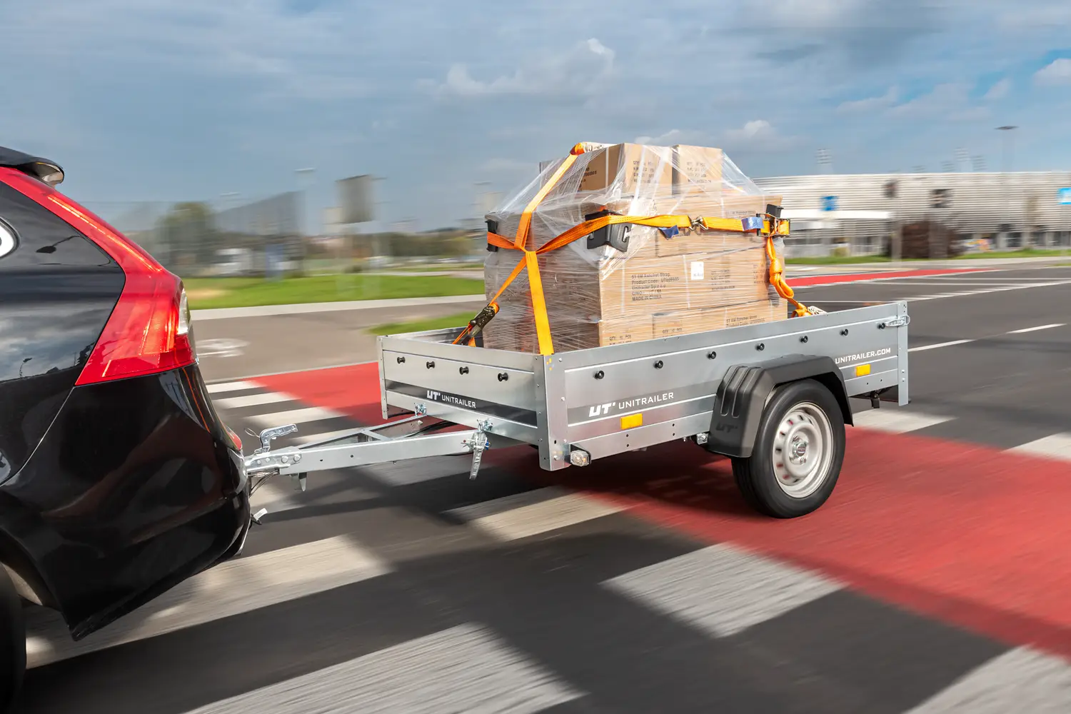 Nákladní přívěs Unitrailer připojený k černému autu, převážející paletu s krabicemi ovinutou stretch fólií a zajištěnou oranžovými upínacími pásy při jízdě po silnici